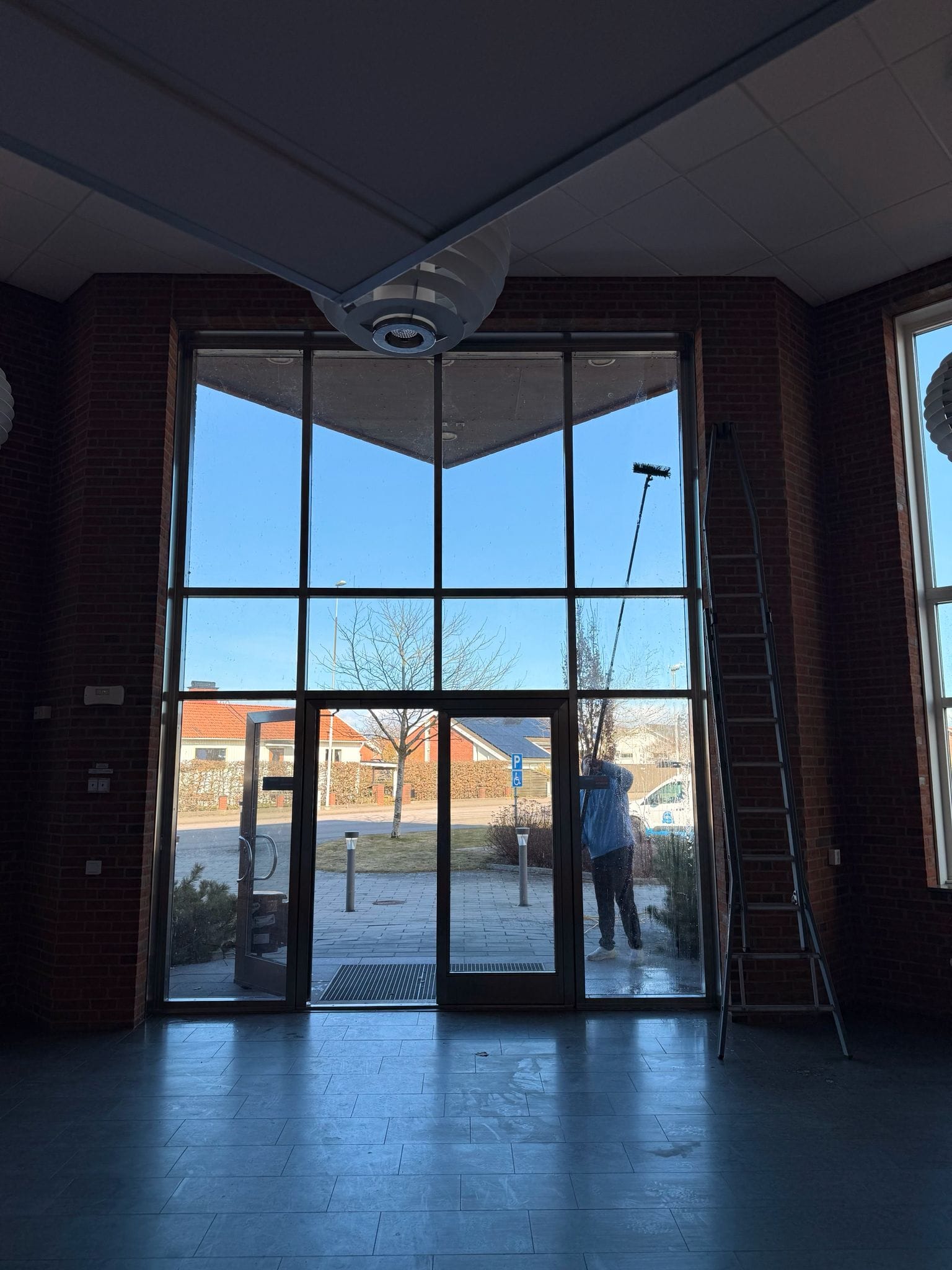 Fönsterputsare rengör ett högt glasparti utifrån med skaft, sett inifrån